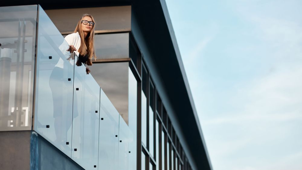 Modern frameless glass balustrade on a high balcony, stand off brackets and aluminium framing, woman leaning on the rail, contemporary building against blue sky.