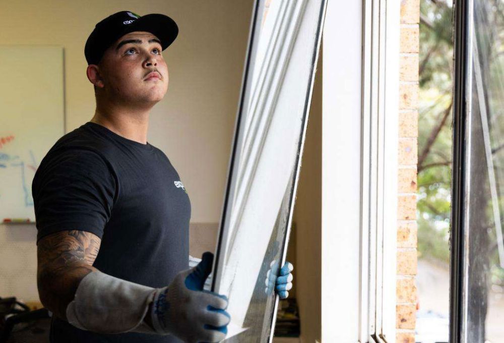 An Express Glass worker wearing gloves is installing a window, looking upwards while holding the window frame. The setting appears to be an interior space with natural light coming through.