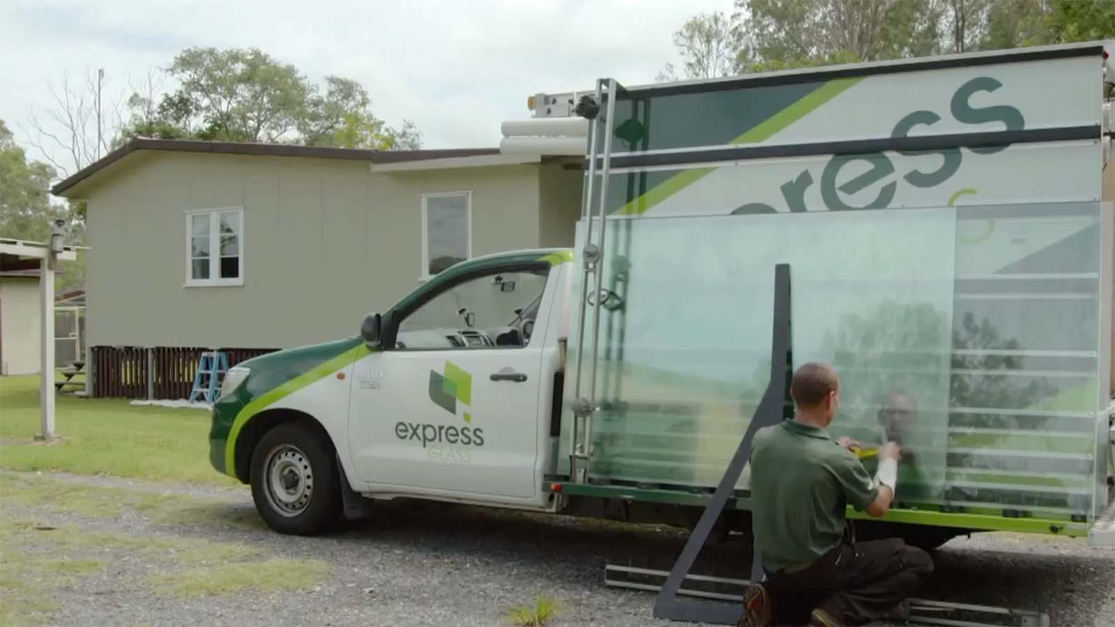 An Express Glass emergency response vehicle with the logo 'express' is parked outside a house. A person is kneeling next to the vehicle, preparing glass panels for installation.