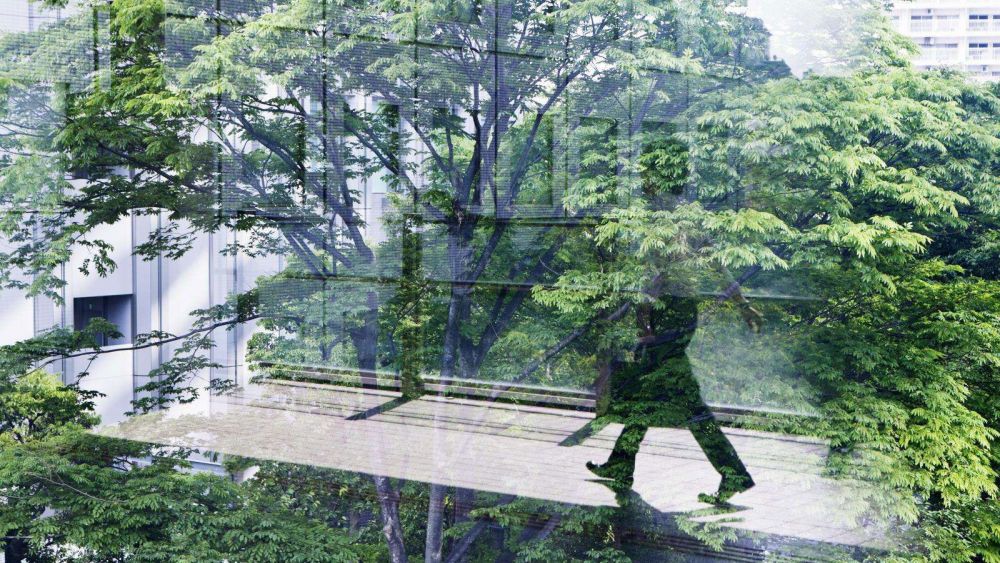 A person walking on a glass bridge between two office towers with tall green trees reflected in the glass, suggesting a blend of nature and urban life.