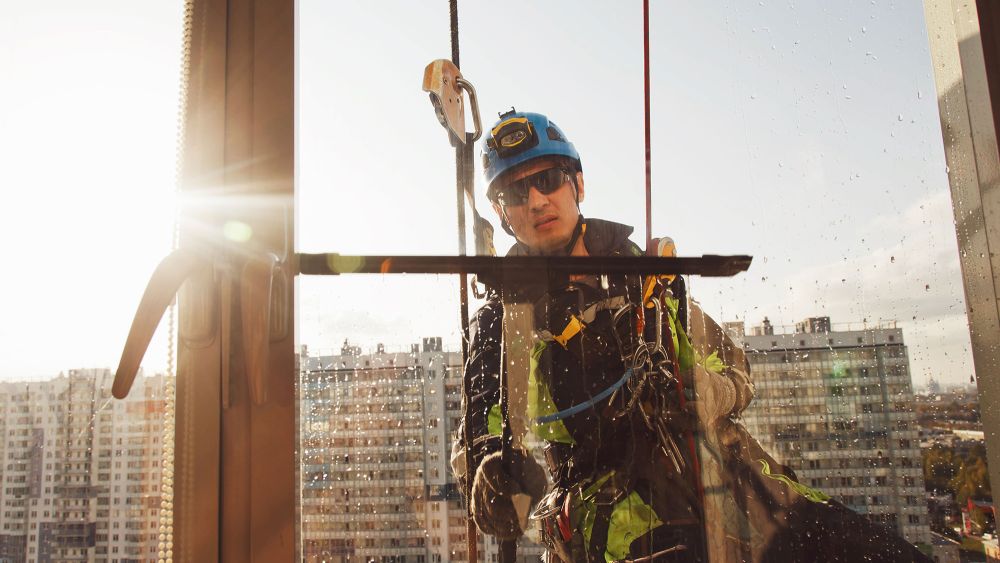 An Express Glass maintenance engineer in safety gear is cleaning a large window on a high-rise building, with the sun shining in the background.