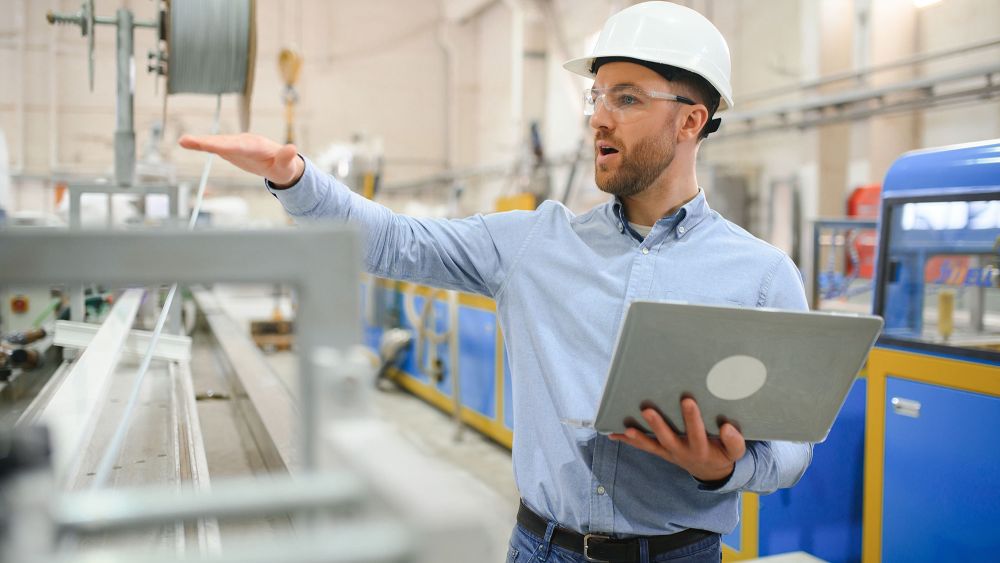 An Express Glass production engineer in a factory wearing a hard hat and safety glasses, gesturing with one hand while holding a laptop in the other, surrounded by industrial aluminium machining equipment.