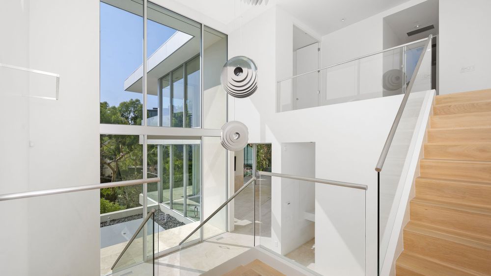 A bright, modern interior featuring a staircase made of light wood with glass balustrades, large glass windows, and a minimalist design with white walls.