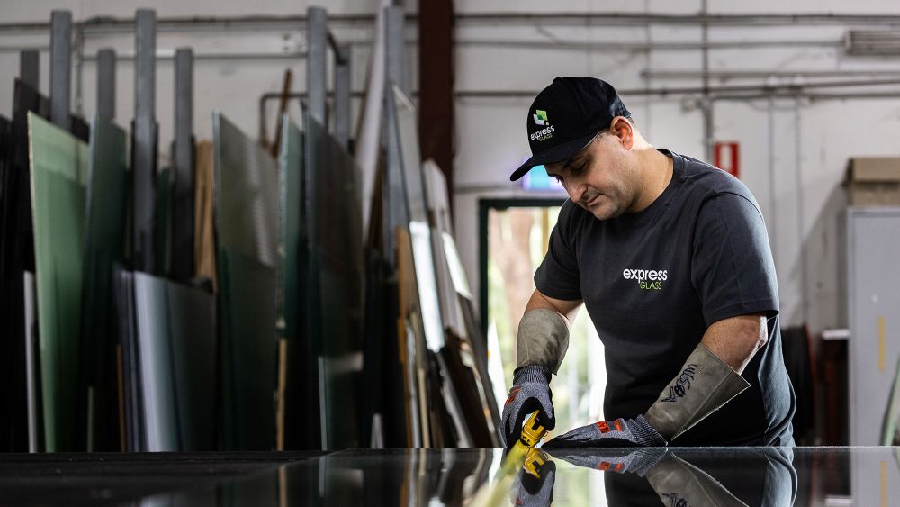 An Express Glass technician in a workshop is measuring a piece of glass on a table, surrounded by various sheets of glass in the background.