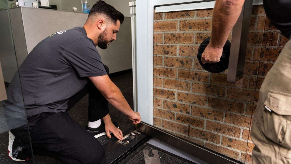 Two Express Glass tradesmen working on repairing a window; one person is kneeling and adjusting a window component, while the other is holding a glass lifting tool.