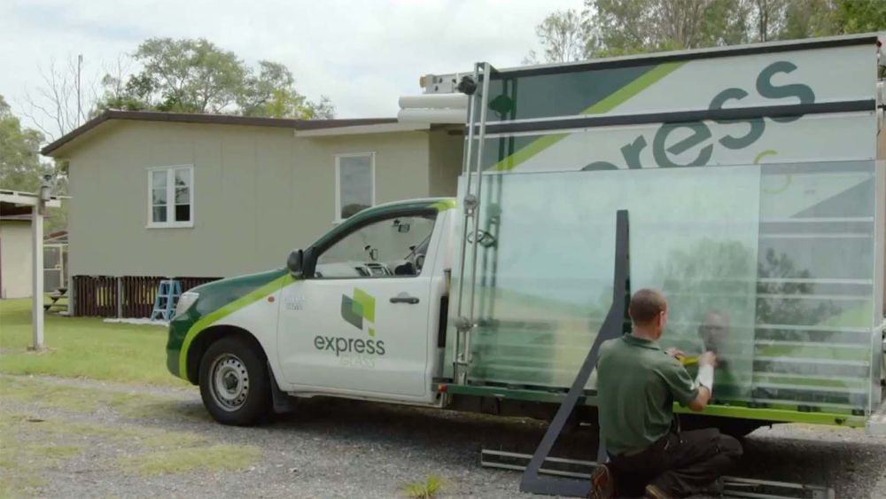 An Express Glass emergency response vehicle with the logo 'express' is parked outside a house. A person is kneeling next to the vehicle, preparing glass panels for installation.