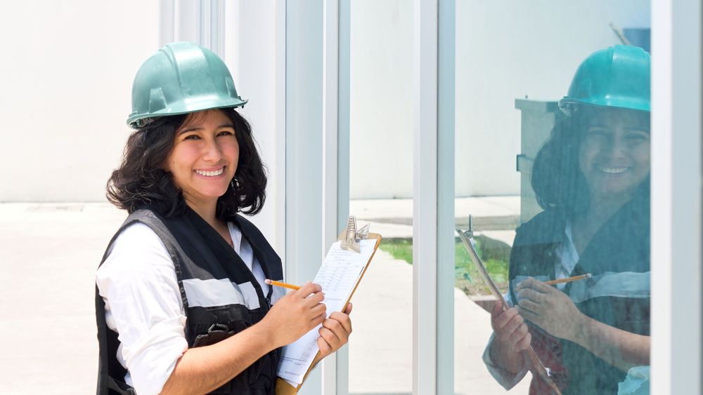 An Express Glass Safety Auditor wearing a green hard hat and a black vest stands next to large glass panels, holding a clipboard and smiling.