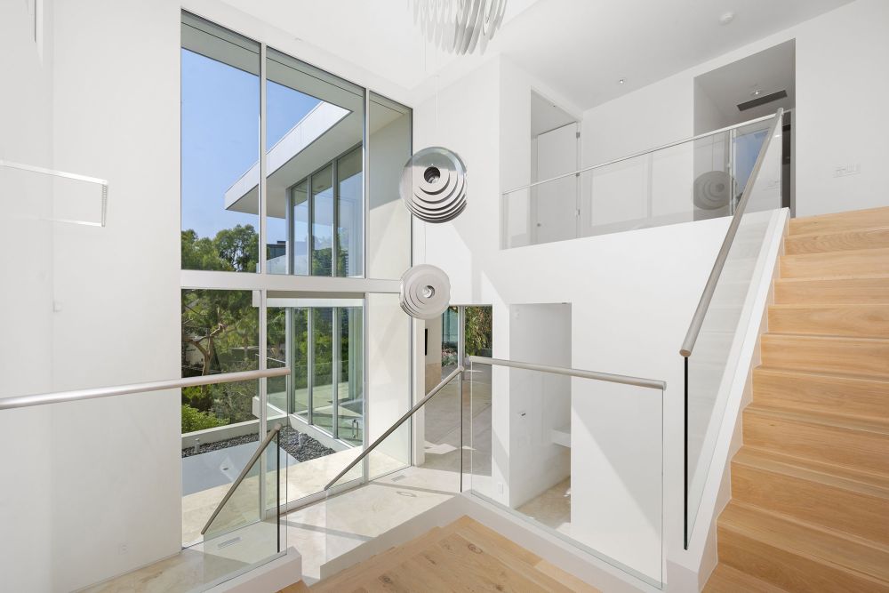 A bright, modern interior featuring a staircase made of light wood with glass balustrades, large glass windows, and a minimalist design with white walls.