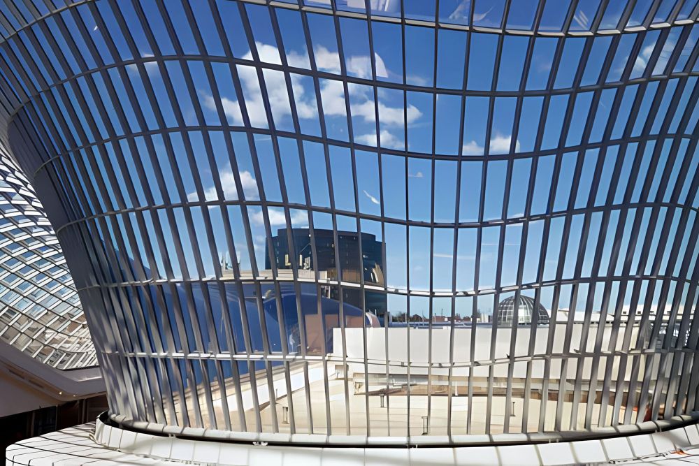 The curved glass structure of Chadstone Shopping Centre reflecting the blue sky and clouds, with a view of nearby buildings in the background.