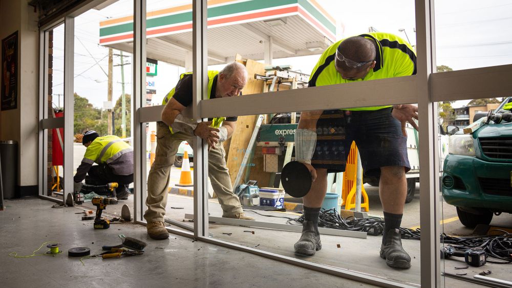 Three Express Glass emergency response workers in high-visibility vests are repairing a 7-Eleven storefront window. Tools and equipment are scattered on the floor, and a 7-Eleven forecourt is visible in the background.