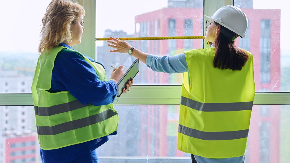 Two women in safety vests are inside a building. One is measuring a window for AS 1288 compliance with a tape measure, while the other is taking notes on a clipboard.