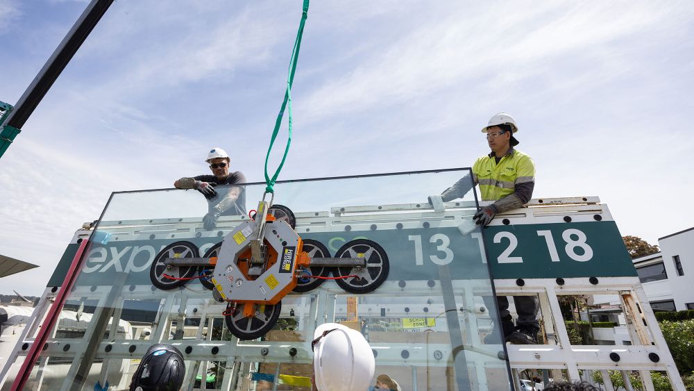 Express Glass workers in hard hats are overseeing the lifting of a large glass panel by a crane. The glass panel is being held by a suction device, and the scene is set outdoors with a clear sky.