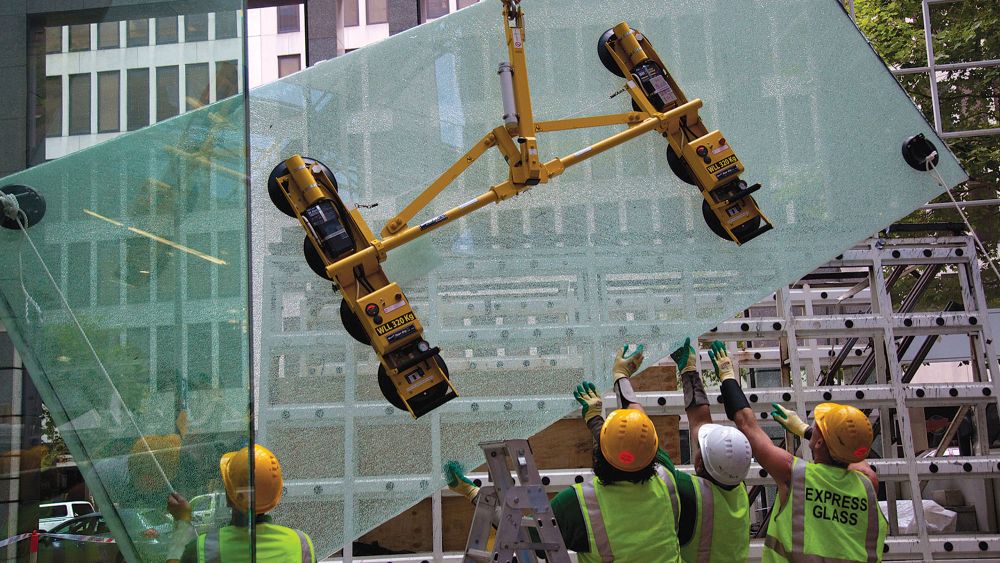 Express Glass workers in safety gear are using a yellow glass-lifting device to install a large glass panel at a building site.