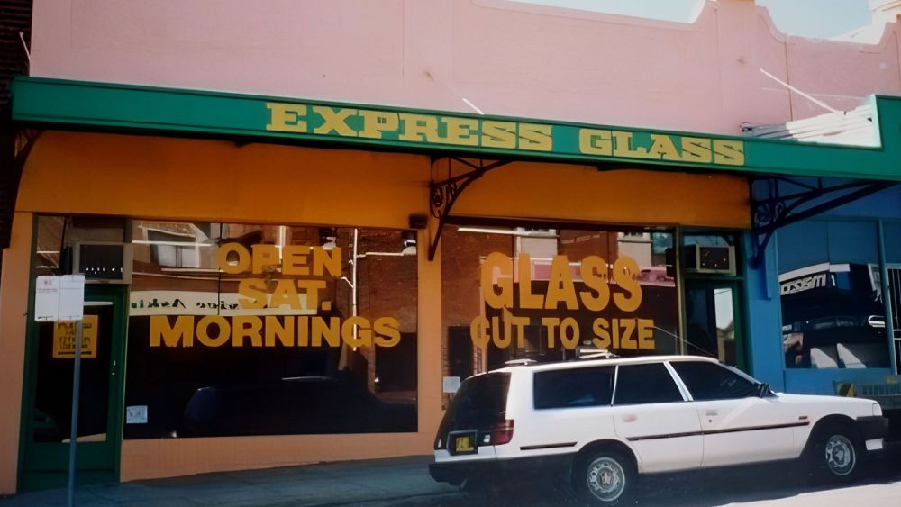 The original storefront for 'Express Glass' with large windows displaying the text 'OPEN MORNINGS' and 'GLASS CUT TO SIZE.' The building has a pink exterior and a green awning. A white car is parked in front.
