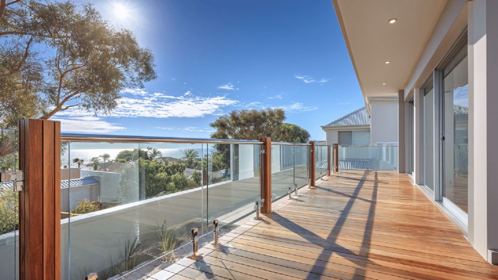 Glass balustrade with timber posts on a wide timber deck of a modern coastal home, clear panels and stainless fittings framing ocean views in bright sun.