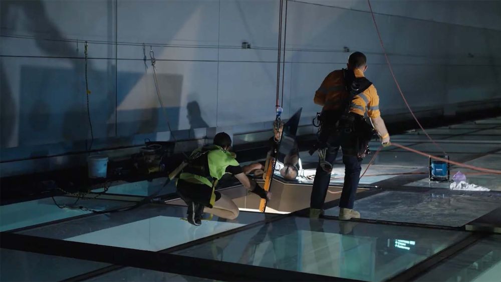 Two Express Glass specialist glaziers working on site at Westfields Bondi Junction, one kneeling and examining equipment while the other stands nearby, with large glass panels visible on the ground.