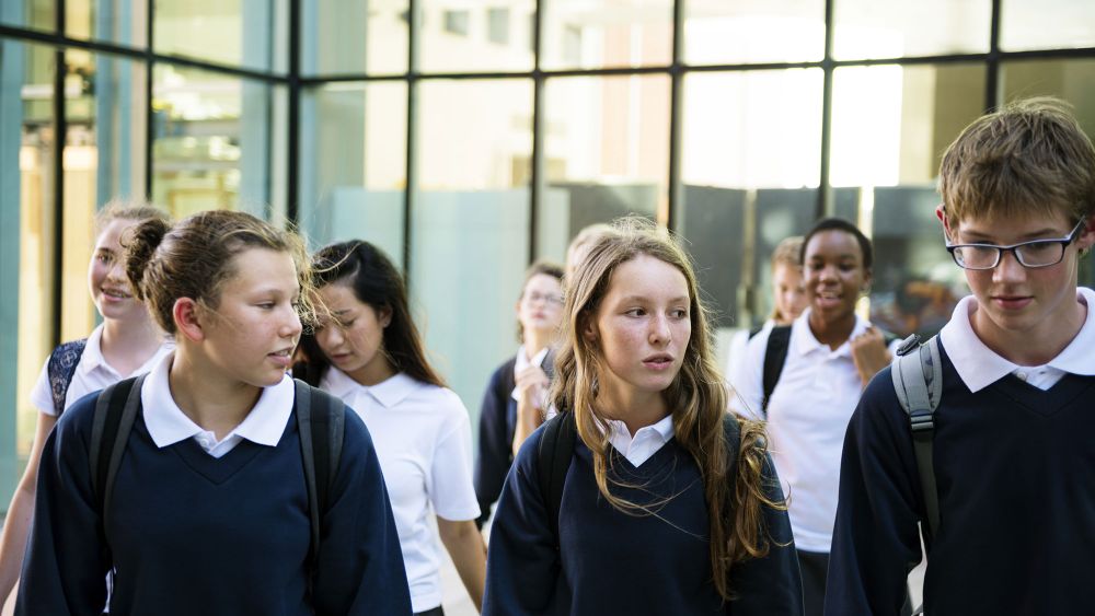 A group of students in school uniforms, consisting of dark sweaters and white collared shirts, walking together in a modern school setting with large glass windows.