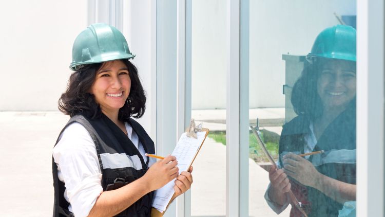 An Express Glass Safety Auditor wearing a green hard hat and a black vest stands next to large glass panels, holding a clipboard and smiling.