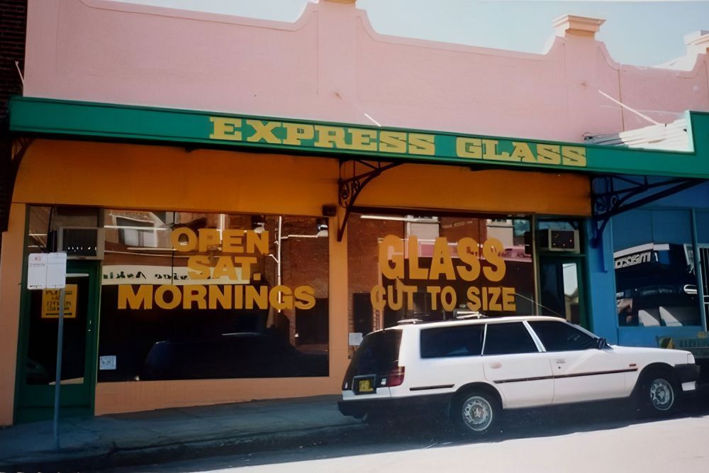 The original storefront for 'Express Glass' with large windows displaying the text 'OPEN MORNINGS' and 'GLASS CUT TO SIZE.' The building has a pink exterior and a green awning. A white car is parked in front.