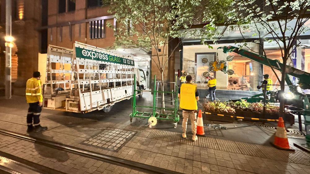 A nighttime street scene with Express Glass emergency response workers in yellow vests near a retail area in George Street Sydney. There are safety cones and a large green trolley, with a retail shop visible in the background.
