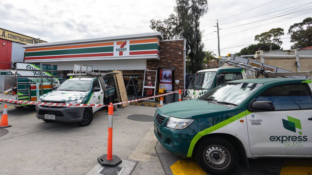 A 7-Eleven convenience store with several Express Glass service trucks outside. The area is marked with traffic cones and caution tape.