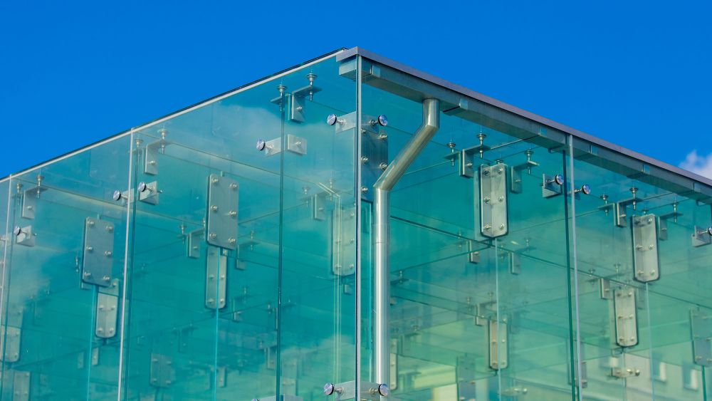 Close-up view of a modern glass building corner, showcasing transparent glass panels and metal fixtures against a blue sky.