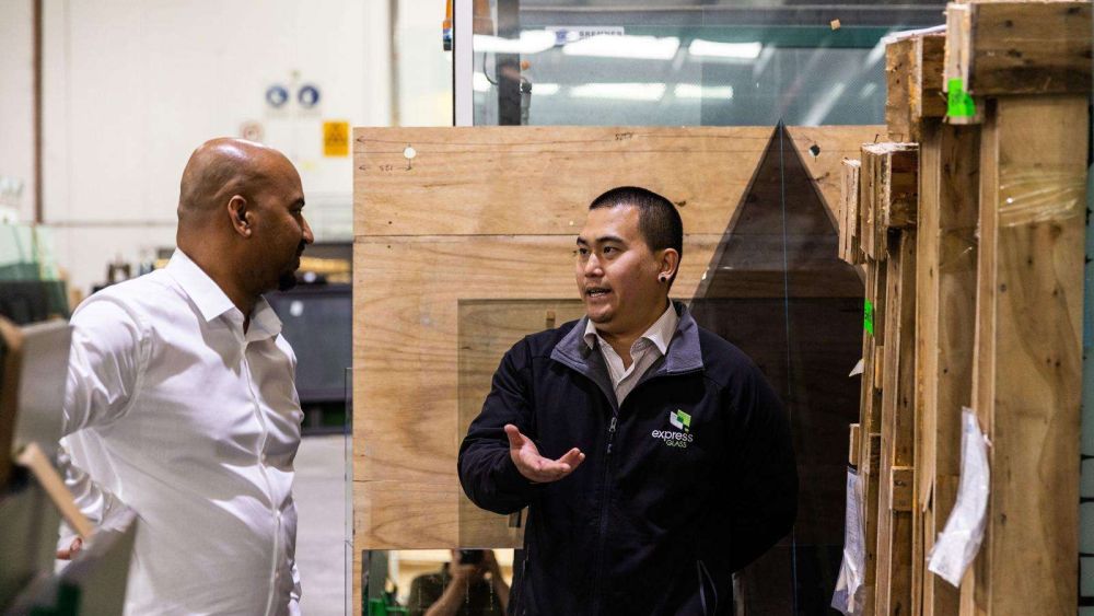 Two Express Glass technicians are talking in a warehouse. One person is wearing a white shirt and the other is in a black jacket with the Express Glass logo on the left breast. They are standing near wooden crates.