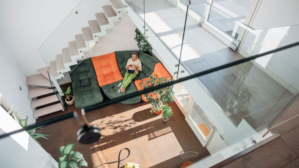 A modern interior with a person sitting on a green sofa, surrounded by plants and natural light, with stairs with glass balustrades visible in the background.