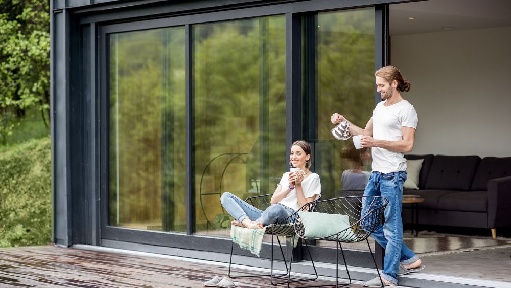 A man stands beside a woman sitting on a chair outside a house with modern residential glazing, both enjoying a leisurely moment. The woman is holding a hot drink, while the man pours another.