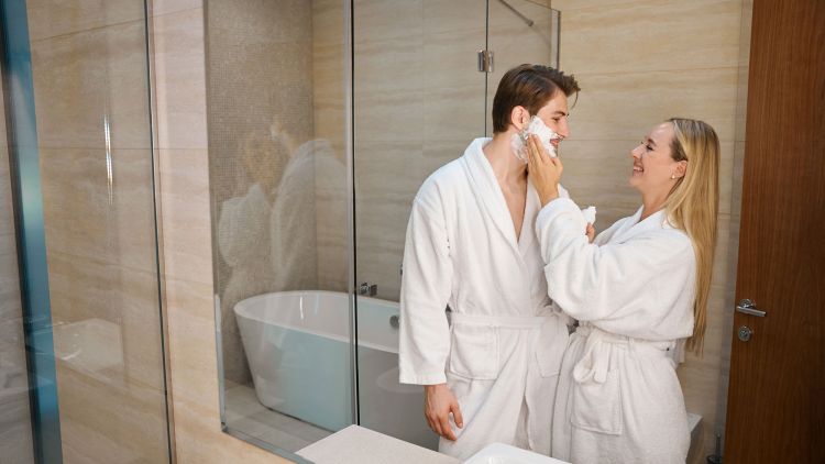 A man and a woman in white bathrobes in a modern bathroom, the woman is applying shaving cream to the man's face, with a bathtub and glass shower visible in the background.
