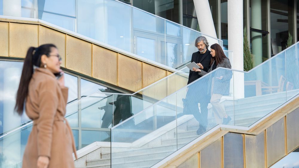 A modern staircase with glass balustrades. Two people are seen walking up the stairs, engaged in conversation, while a third person in a long coat walks past them, talking on a phone.