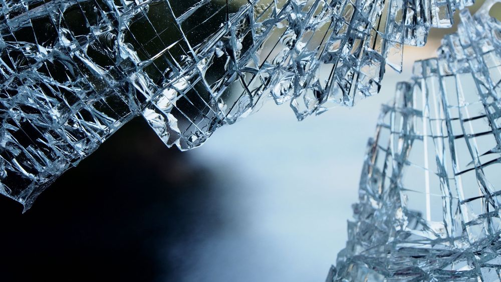 Close-up of broken glass with sharp edges, reflecting light, against a blurred dark background.