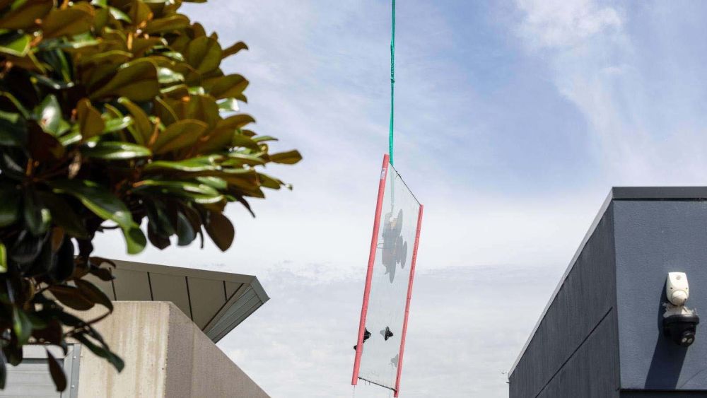 A large glass panel is suspended in the air by a green rope, with two buildings visible on either side and a cloudy sky above.