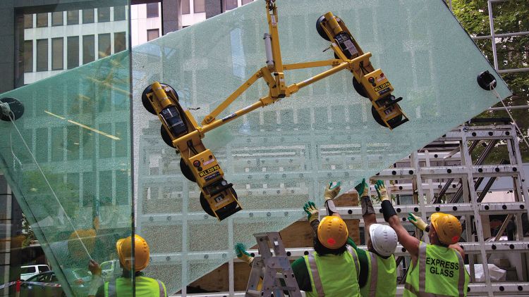Express Glass workers in safety gear are using a yellow glass-lifting device to install a large glass panel at a building site.