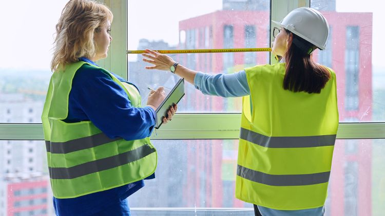 Two women in safety vests are inside a building. One is measuring a window for AS 1288 compliance with a tape measure, while the other is taking notes on a clipboard.