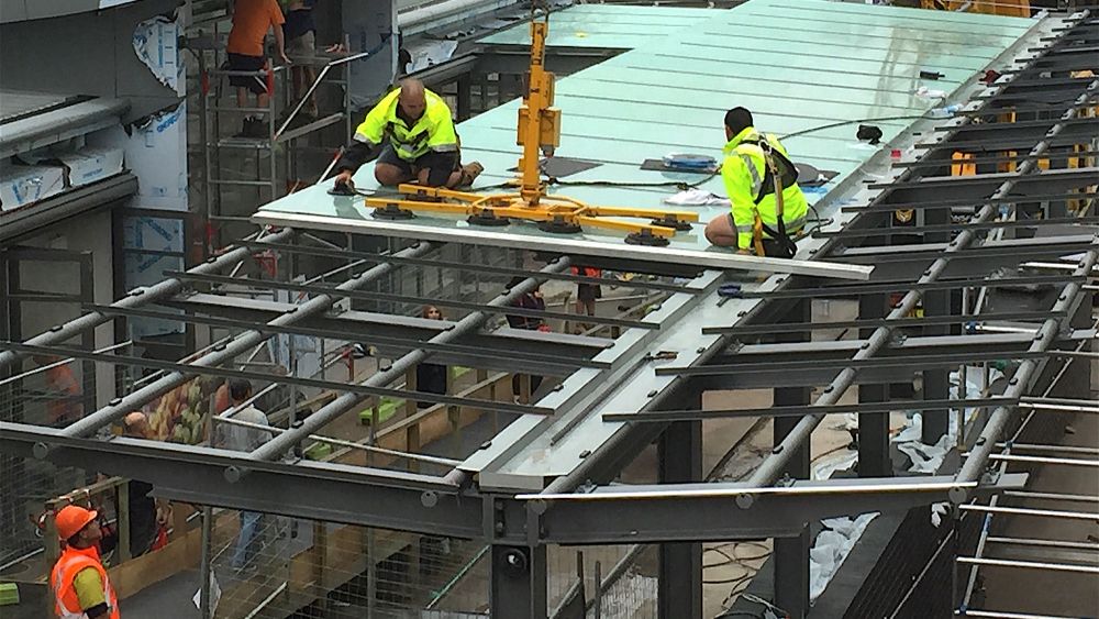 Express Glass workers in high-visibility clothing are working on a steel framework, placing a large glass panel on top of a structure at Ashfield Mall. The scene shows cranes and scaffolding, indicating an active construction site.