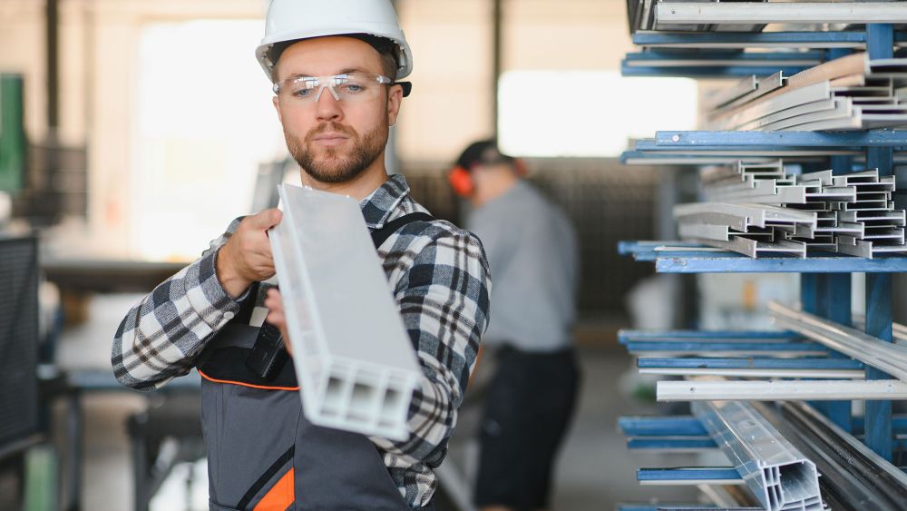 An Express Glass production engineer wearing a hard hat and safety glasses holds a long piece of aluminium in a manufacturing setting, with shelves of aluminium material in the background.