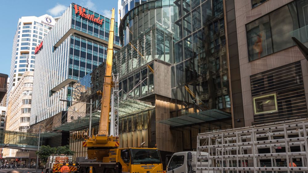 A busy city street with a large yellow construction crane, trucks, and modern glass buildings. The crane is working to lift a large glass pane up to the facade of a commercial building.