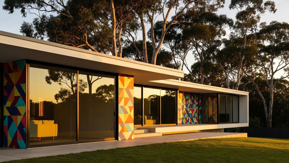 A modern house with colourful geometric patterns on its walls, set in a natural environment with trees, during sunset. The window film on the glass is reflecting distant trees and the sky.