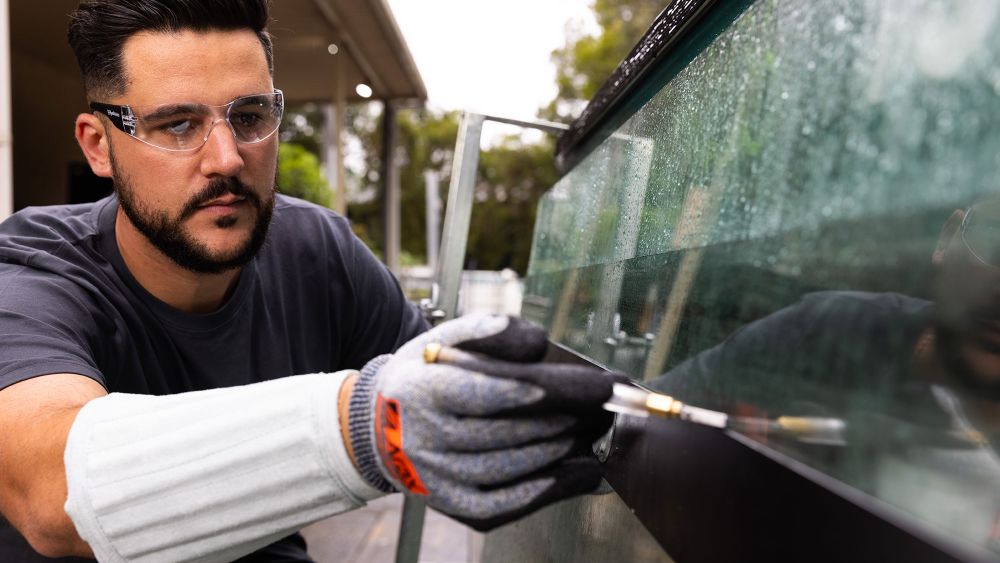 An Express Glass worker wearing safety glasses and gloves is carefully a replacement glass pane off a truck, indicating he is repairing a window.