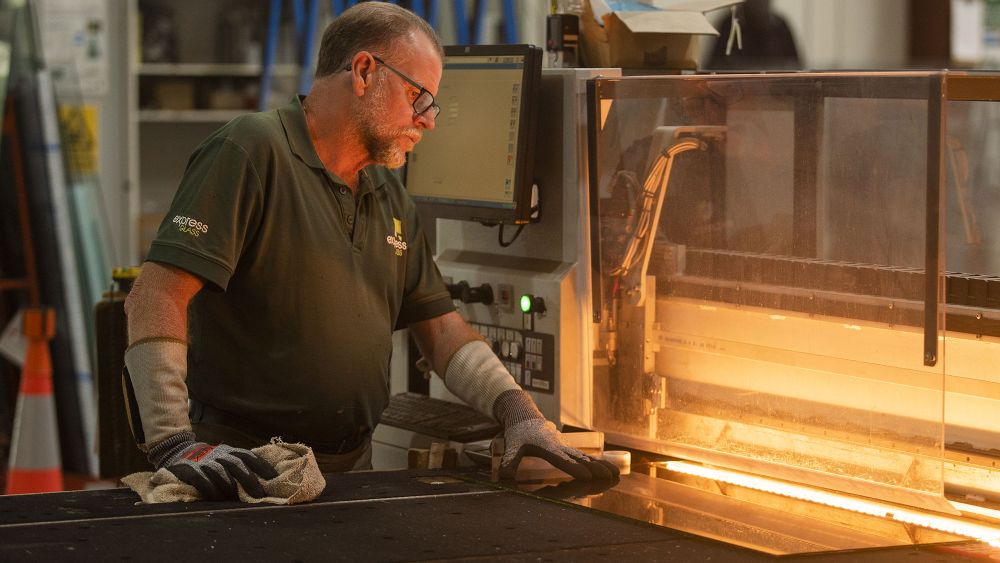 An Express Glass expert glazier with glasses and gloves operates a machine in a workshop, with a glowing element indicating the machine is in use.