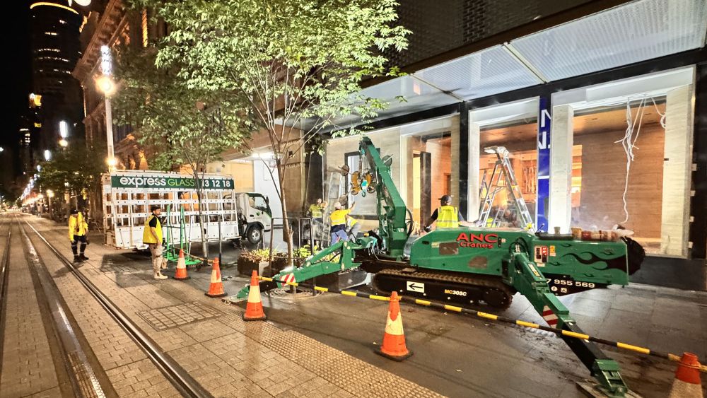 A construction site at night with several Express Glass emergency response workers in safety vests, a green crane, and orange safety cones. The background shows a retail shop with large windows.