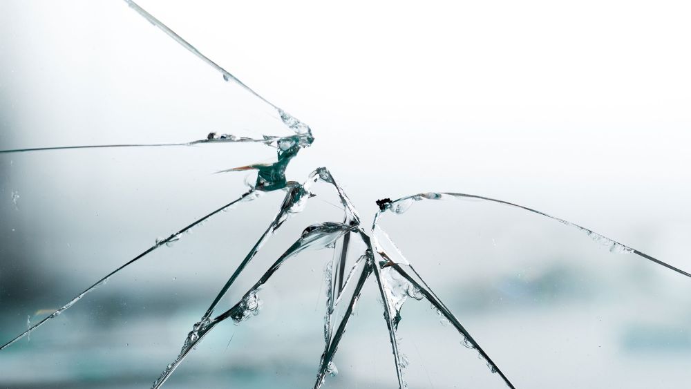 Close-up of a cracked glass surface with multiple visible cracks radiating from a central point, set against a blurred background.