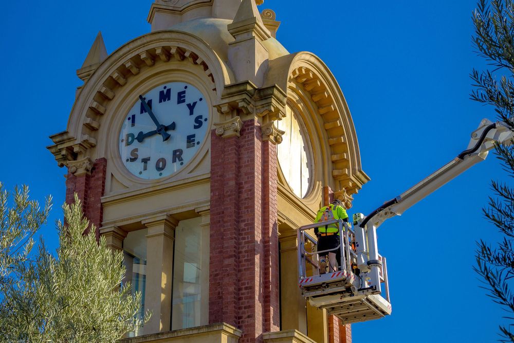An Express Glass worker on a boom lift is repairing the famous Dimmeys clock on a tower, set against a clear blue sky.