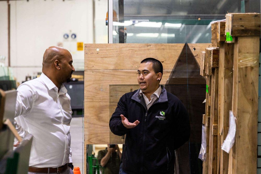 Two Express Glass technicians are talking in a warehouse. One person is wearing a white shirt and the other is in a black jacket with the Express Glass logo on the left breast. They are standing near wooden crates.