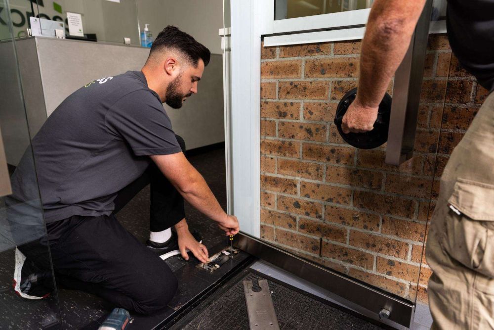 Two Express Glass tradesmen working on repairing a window; one person is kneeling and adjusting a window component, while the other is holding a glass lifting tool.
