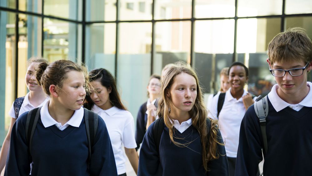 A group of students in school uniforms, consisting of dark sweaters and white collared shirts, walking together in a modern school setting with large glass windows.