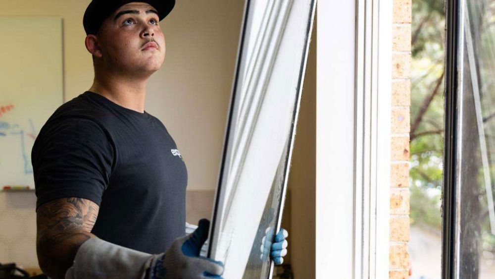An Express Glass worker wearing gloves is installing a window, looking upwards while holding the window frame. The setting appears to be an interior space with natural light coming through.