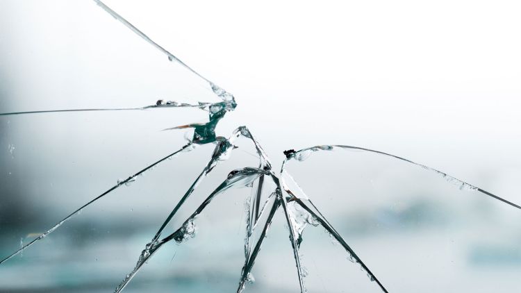 Close-up of a cracked glass surface with multiple visible cracks radiating from a central point, set against a blurred background.