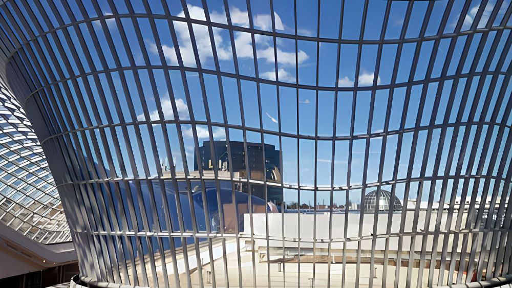 The curved glass structure of Chadstone Shopping Centre reflecting the blue sky and clouds, with a view of nearby buildings in the background.